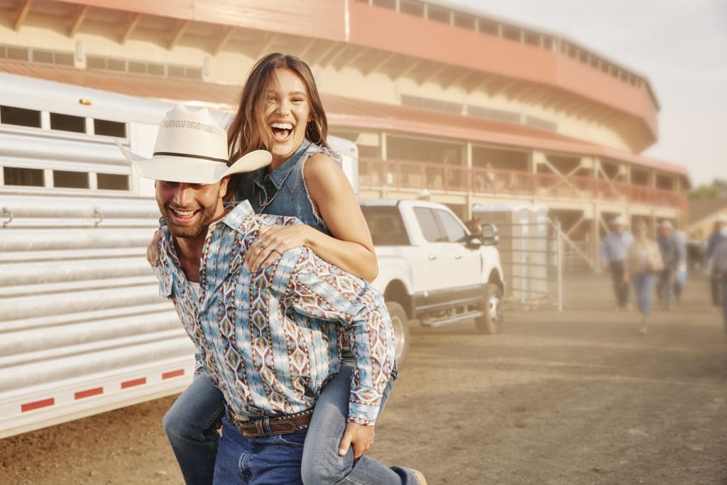 Cowboy joyfully carries a smiling girl on his back, surrounded by a rustic outdoor rodeo setting. A truck and horse trailer are parked on a dusty lot, evoking the spirit of rodeo adventure.