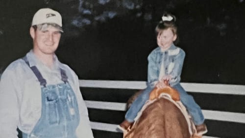 A nostalgic photo of a young Madison Davis sitting on a horse, smiling, with her dad standing beside her. Madison is dressed in a denim outfit, and her dad is wearing overalls and a cap. The image captures a heartwarming moment, emphasizing the strong bond between father and daughter in a rural setting, celebrating the early beginnings of Madison's cowgirl journey.