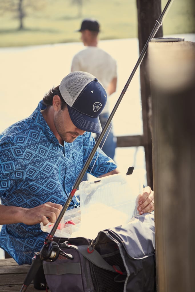 Two men fishing with one actively reeling in his line, while another man in a blue western shirt and ball cap prepares a new lure for his fishing pole.