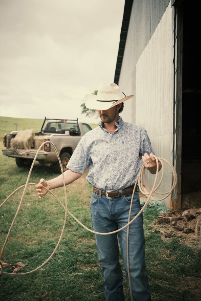 Man in western attire and cowboy hat honing roping skills outdoors, near a barn and pickup truck.