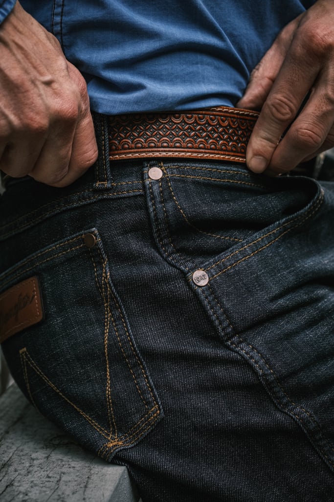 Man wearing Wrangler jeans, sporting a brown leather belt, and donning a blue shirt.