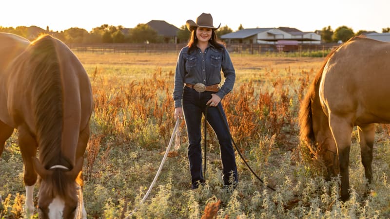 Madison Davis, dressed in a denim shirt and cowboy hat, stands confidently in a field with her two horses. The image captures the essence of a cowgirl’s life, showcasing Madison’s connection with her horses and the rural setting around her.