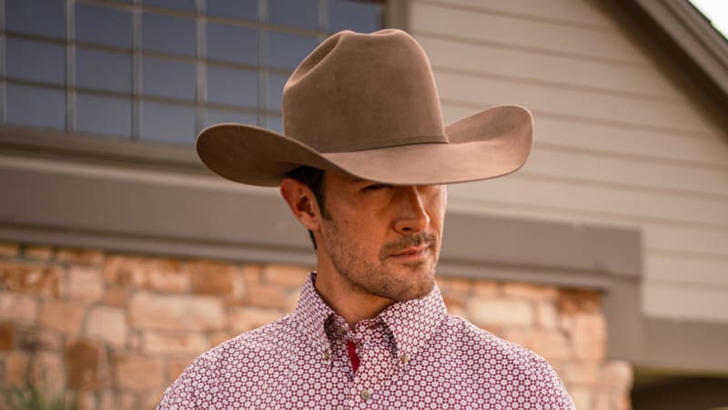 A close-up of a cowboy wearing a wide-brimmed brown felt hat and a patterned button-up shirt. The cowboy has a serious expression, and the background features a stone and wood exterior, possibly of a house or barn, with large windows visible. The setting gives a rustic and western feel to the image.