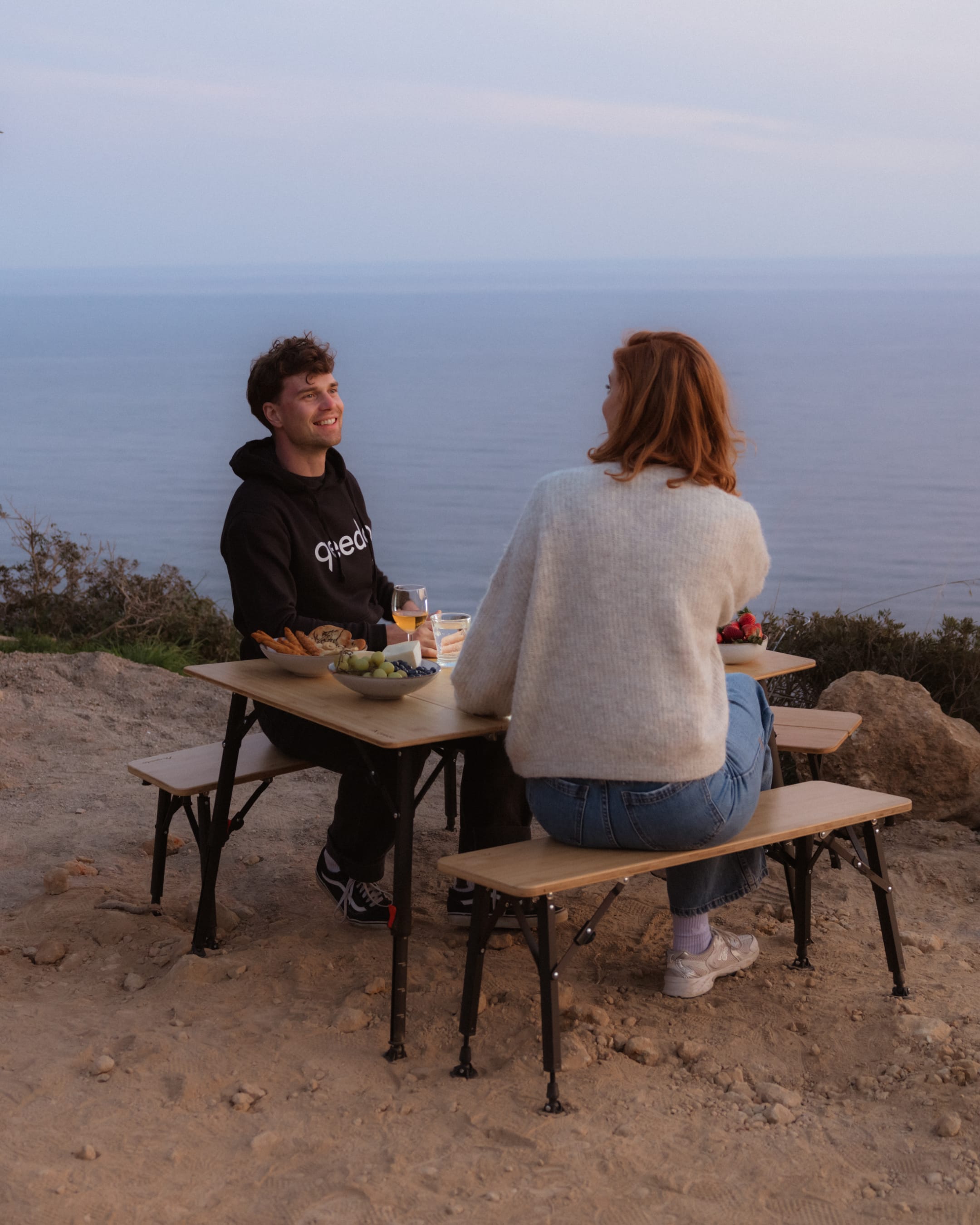 Mann und Frau sitzen auf der klappbaren Sitzbank qeedo Benny an einem Quick Kimmy Campingtisch mit Blick aufs Meer.