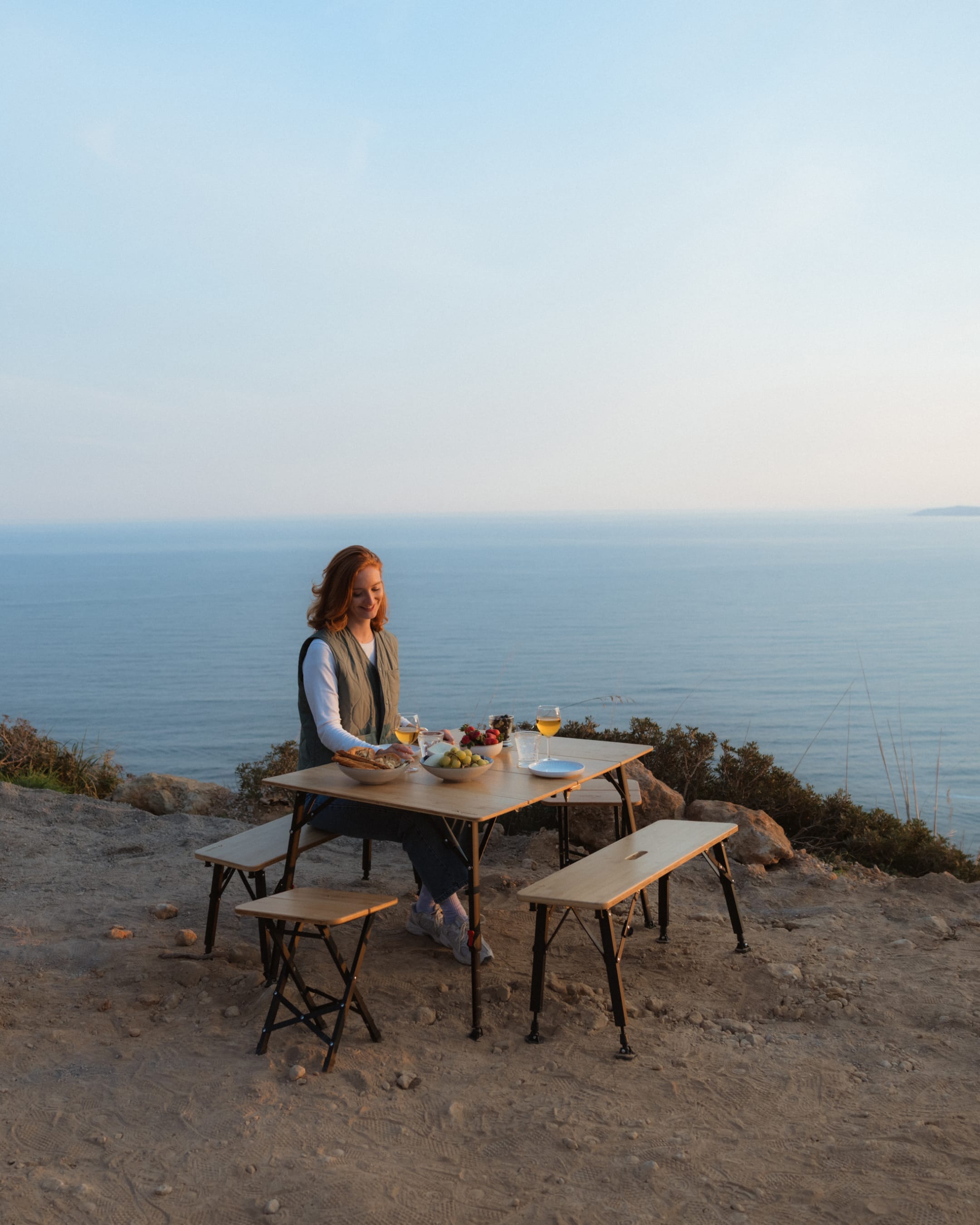 Frau sitzt auf der qeedo Benny Sitzbank am gedecktem qeedo Kimmy Campingtisch mit Aussicht auf das Meer.