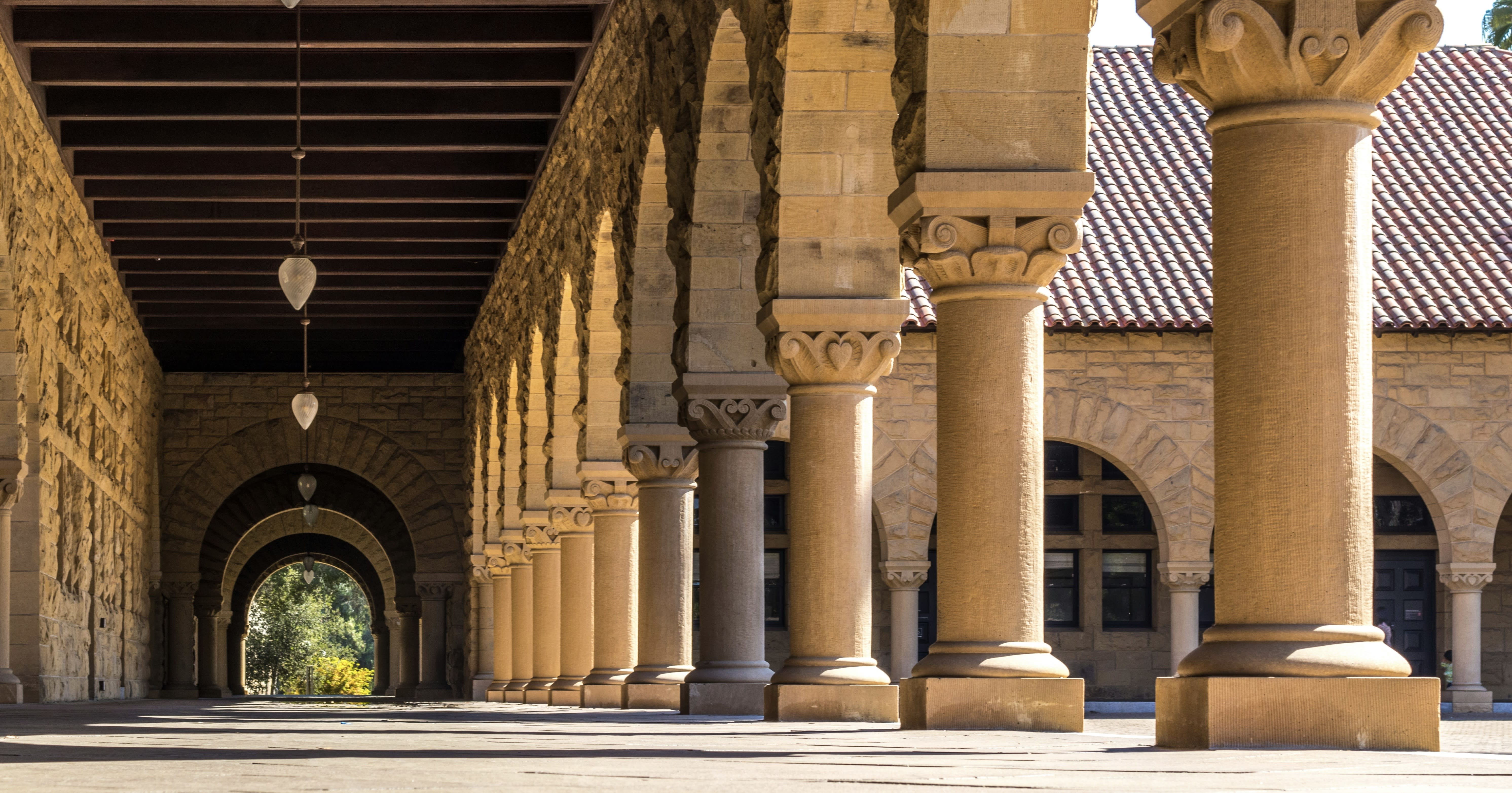 A sunlit colonnade at Stanford University featuring sandstone arches, carved columns, and a tiled roof, creating a calm and tranquil atmosphere.
