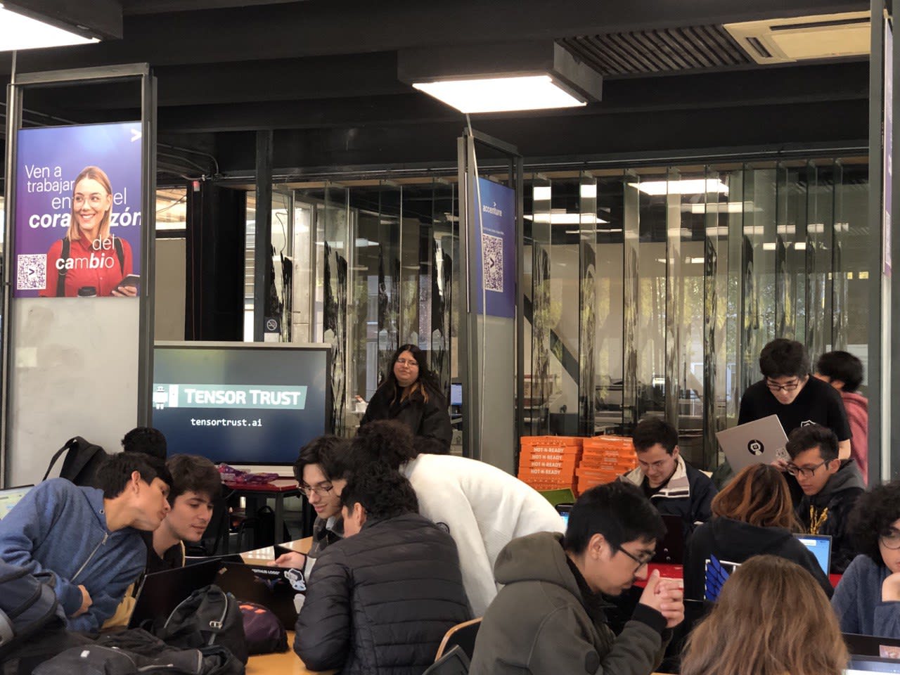 Photo of young people sitting in groups in a crowded university study room. All of them are either looking at their laptops or looking at someone else's laptop over their shoulder. In the background, one can see a TV displaying tensortrust.ai in monospace font and two stacks of still unopened pizza boxes.