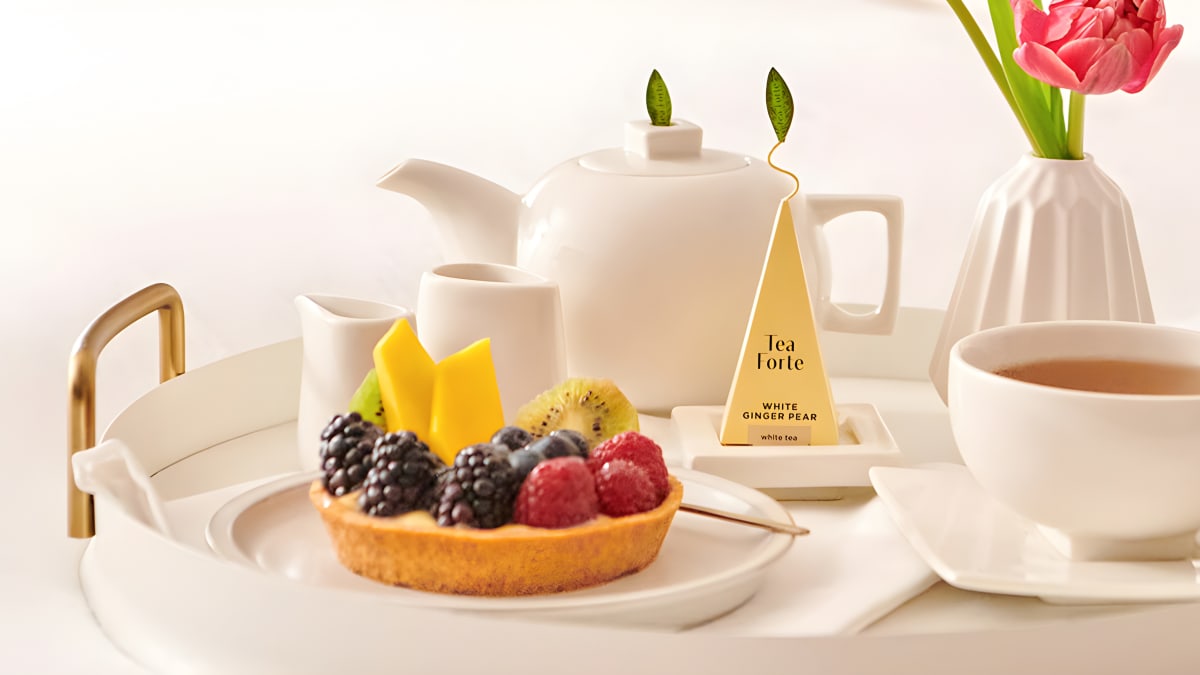 Tea Forte pyramid infuser with its leaf on a white tea tray with a teapot and fruit tart