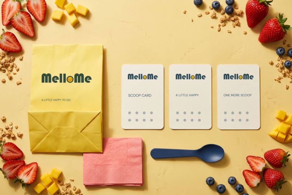 A vibrant flat lay showing MelloMe packaging collaterals including a yellow paper bag, three dotted loyalty "scoop cards," a blue spoon, and pink napkins, surrounded by fresh strawberries, blueberries, mango chunks, and granola.