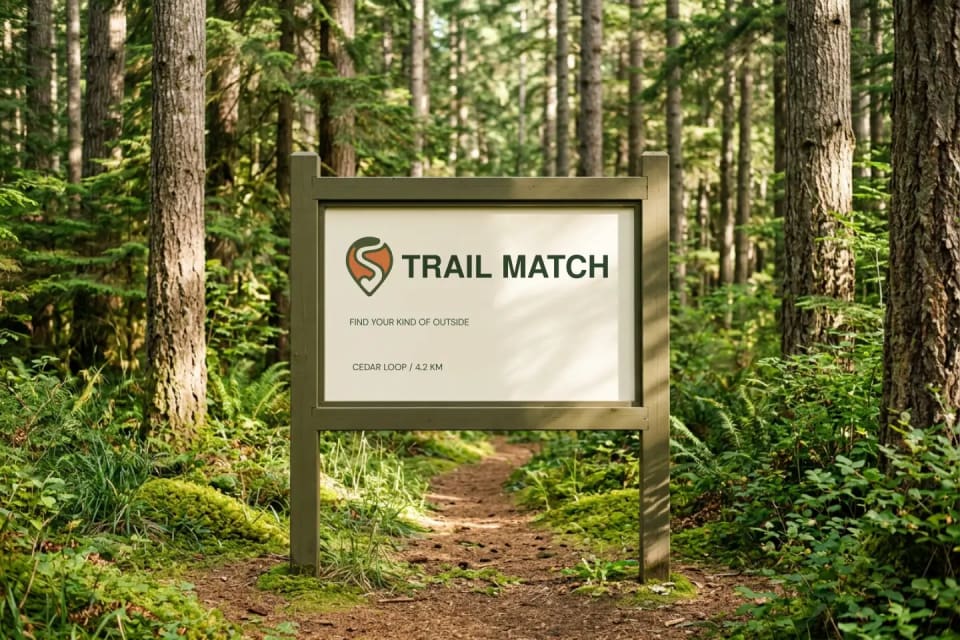 An environmental branding mockup showing a wooden Trail Match sign reading "Cedar Loop / 4.2 KM" placed along a sunlit, narrow dirt path in a dense green forest.