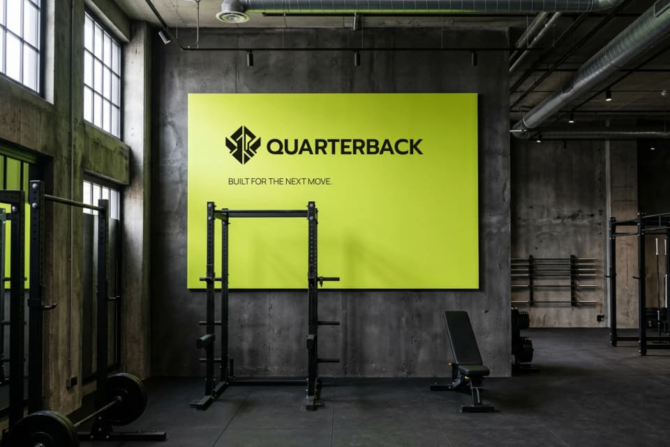 A modern, industrial gym environment for Quarterback, featuring a large, vibrant Signal Lime sign with the brand logo and the tagline "BUILT FOR THE NEXT MOVE." mounted on a dark concrete wall behind workout equipment.