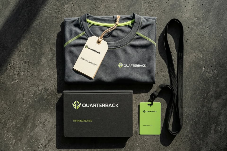A flat lay mockup of Quarterback training gear on a concrete texture, featuring a dark grey athletic shirt with neon green stitching, a training notes box, a branded tag, and a neon green member ID badge on a lanyard.