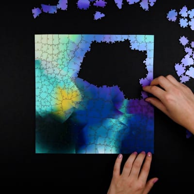 Top-down photo of two hands solving a square jigsaw puzzle. The jigsaws pieces are random polygon shapes of various sizes, and the pattern is abstract with purple, blue, and yellow accents.