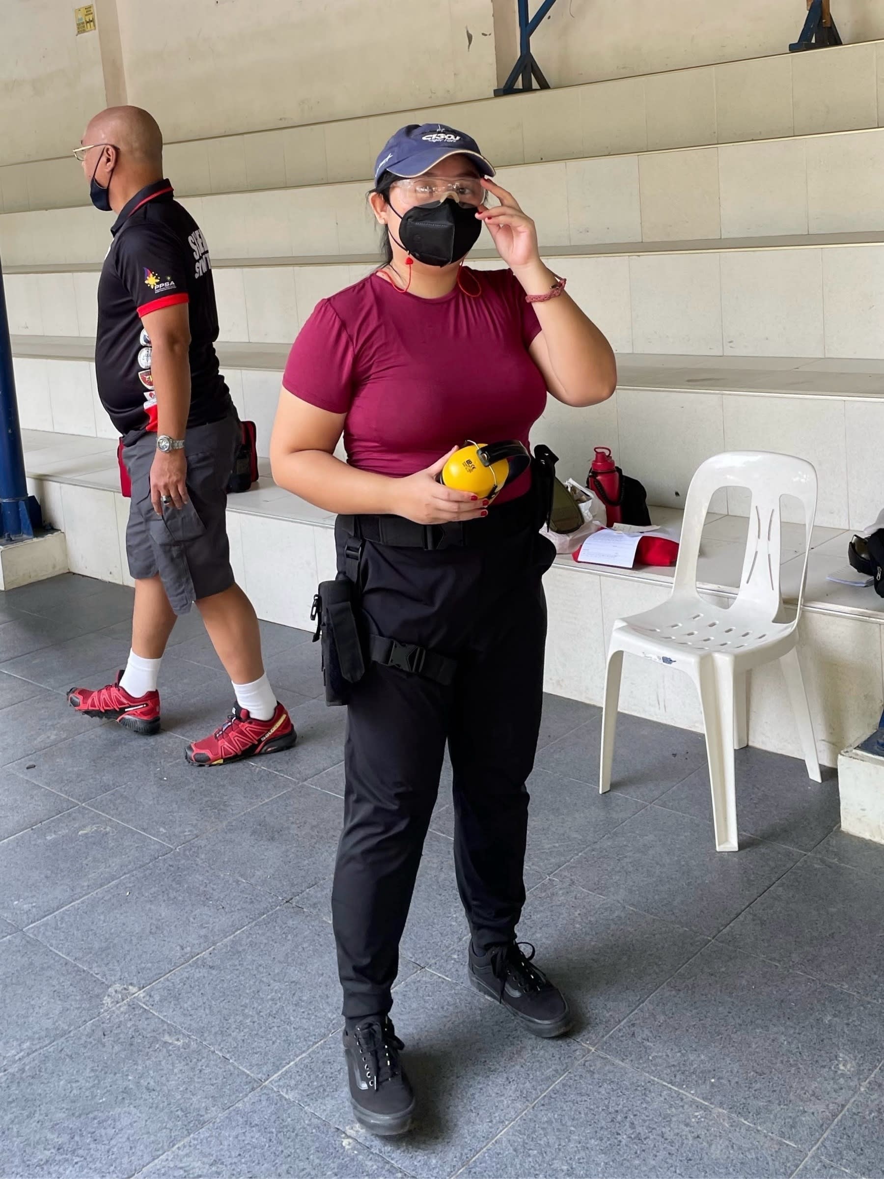 Chi wearing a cap, with her hair tied in a ponytail and swept to the side of her shoulder, a maroon fitted top, and black pants and black shoes. Around her belt and thigh is the belt holster for carrying guns. She's holding onto the noise-dampening headset and she has protective eyeglasses on, and also has a mask on.