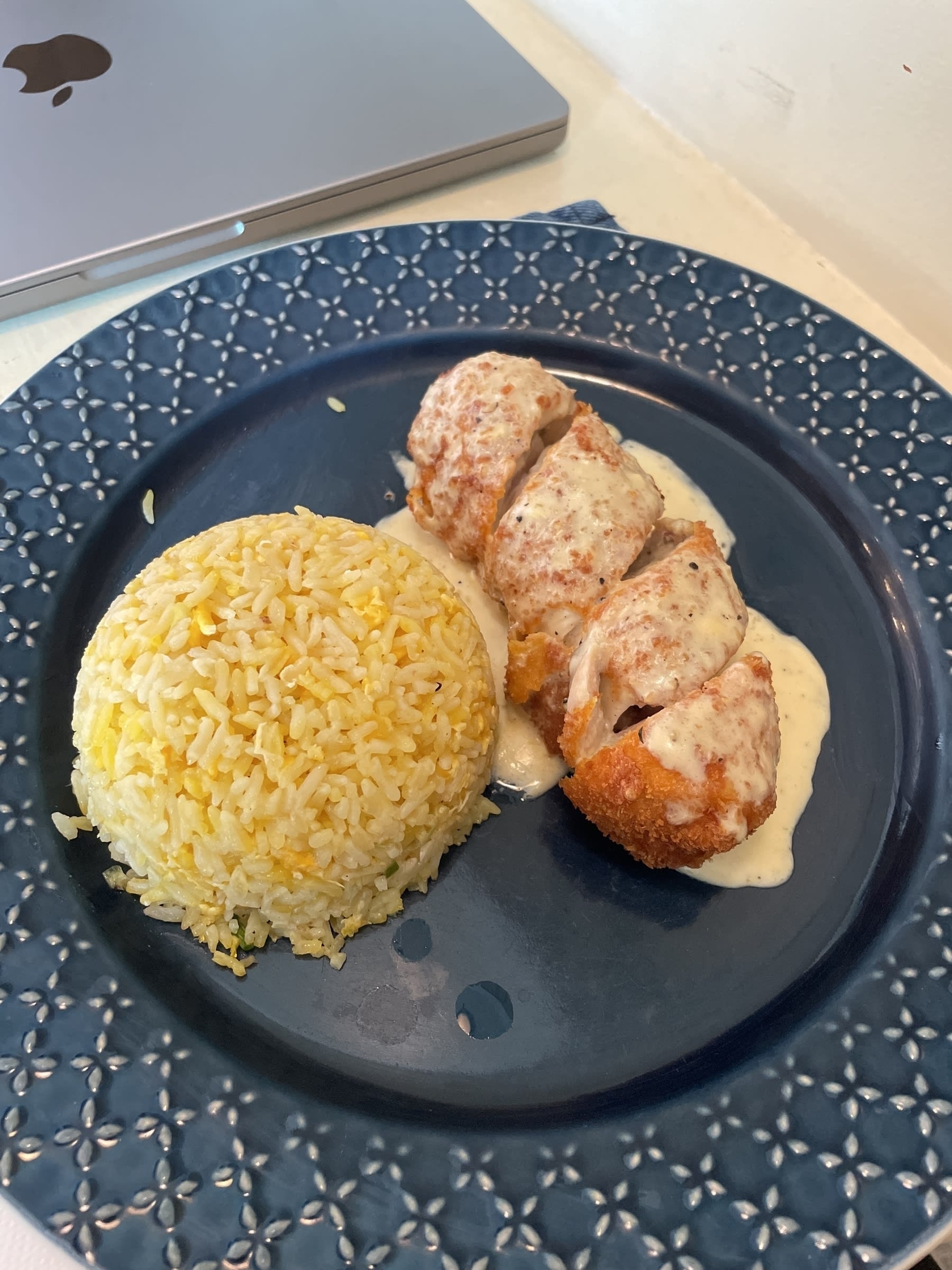 Chicken cordon bleu meal with buttered rice placed on a blue plate. A closed Macbook is seen on the upper left corner of the photo.