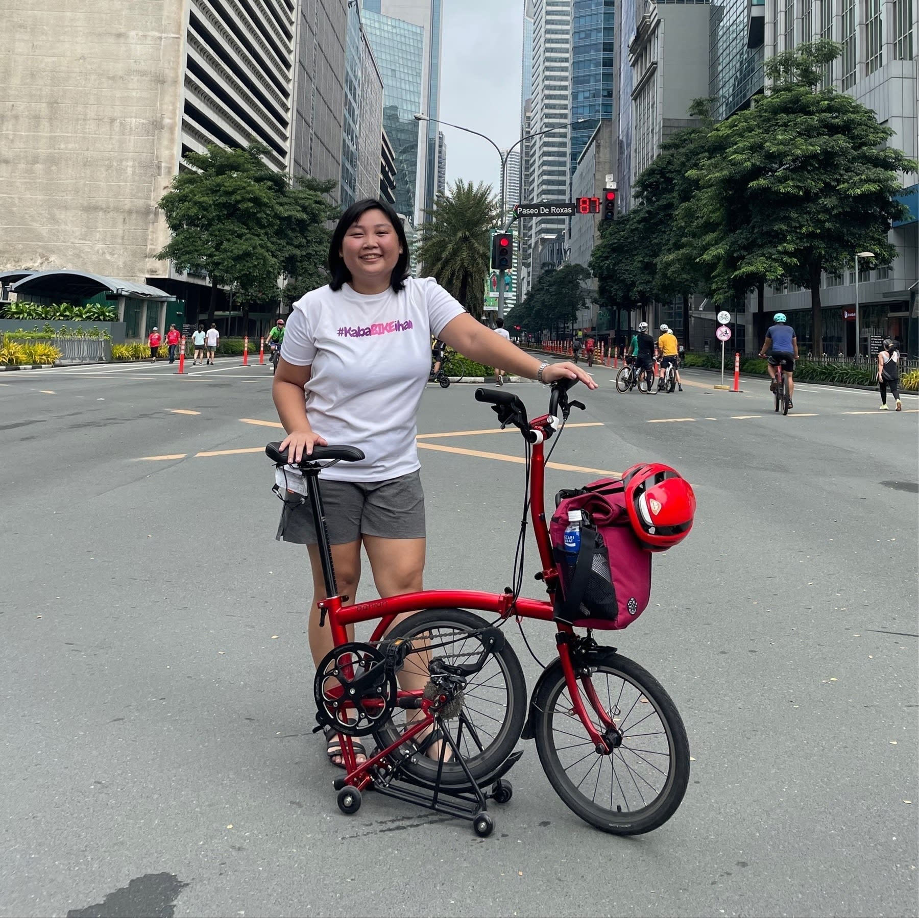 Chi smiling and posing with her red Banian trifold bike, which is partially folded, while in the middle of Ayala Avenue because it's a Car-Free Sunday morning event. She is wearing a white t-shirt that has the text, “#KabaBIKEihan” printed, and gray shorts and black sandals. On her bike, her red GoFar front mounted bag is attached, and her red Lumos helmet is also strapped to her bag.