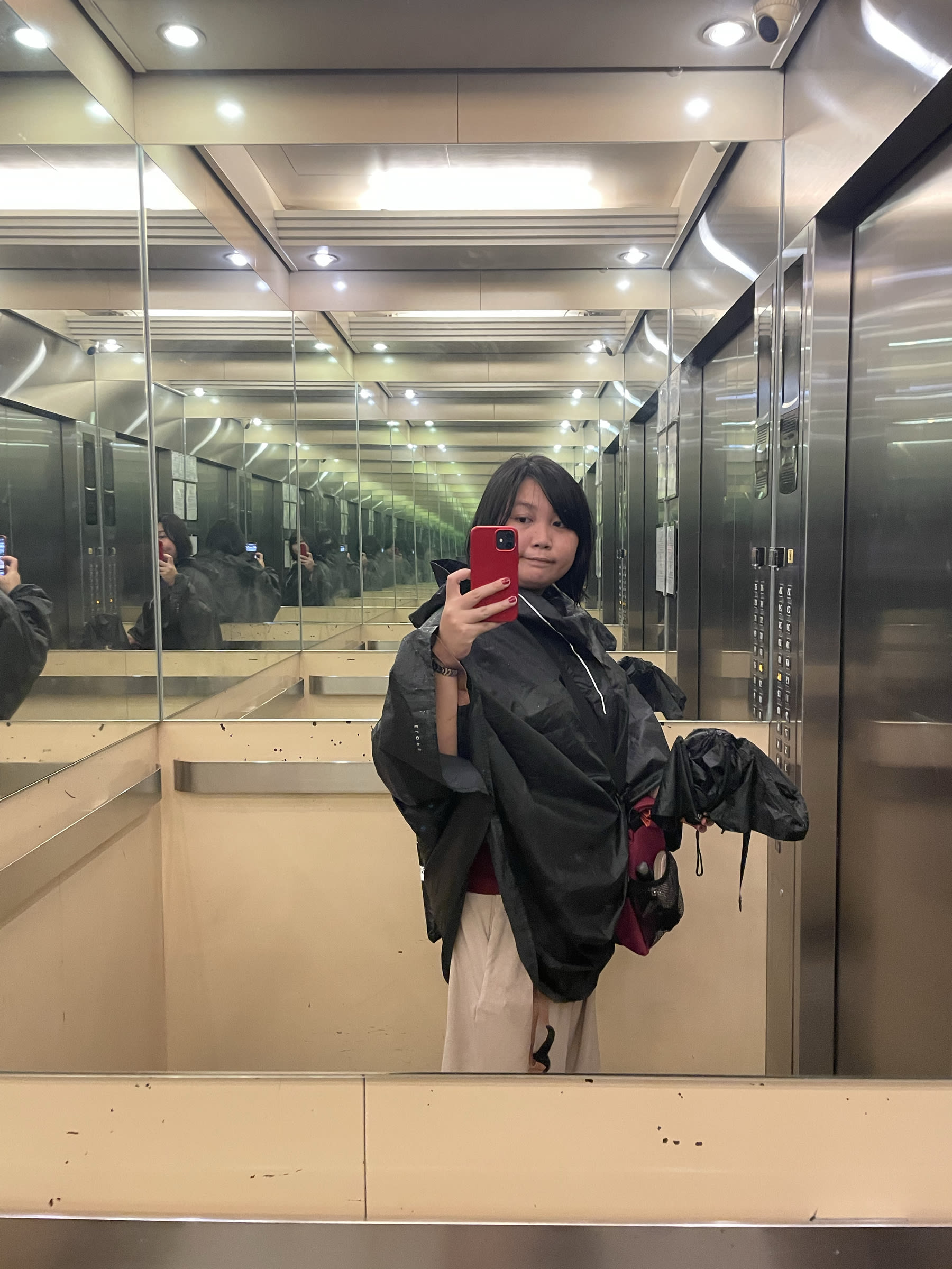 mirror photo of Chi in their condominium's elevator. She is seen wearing her bike poncho over her work clothes. Her bike bag is in sling mode and set to her left, and in her left hand she is also seen holding a wet folded umbrella.