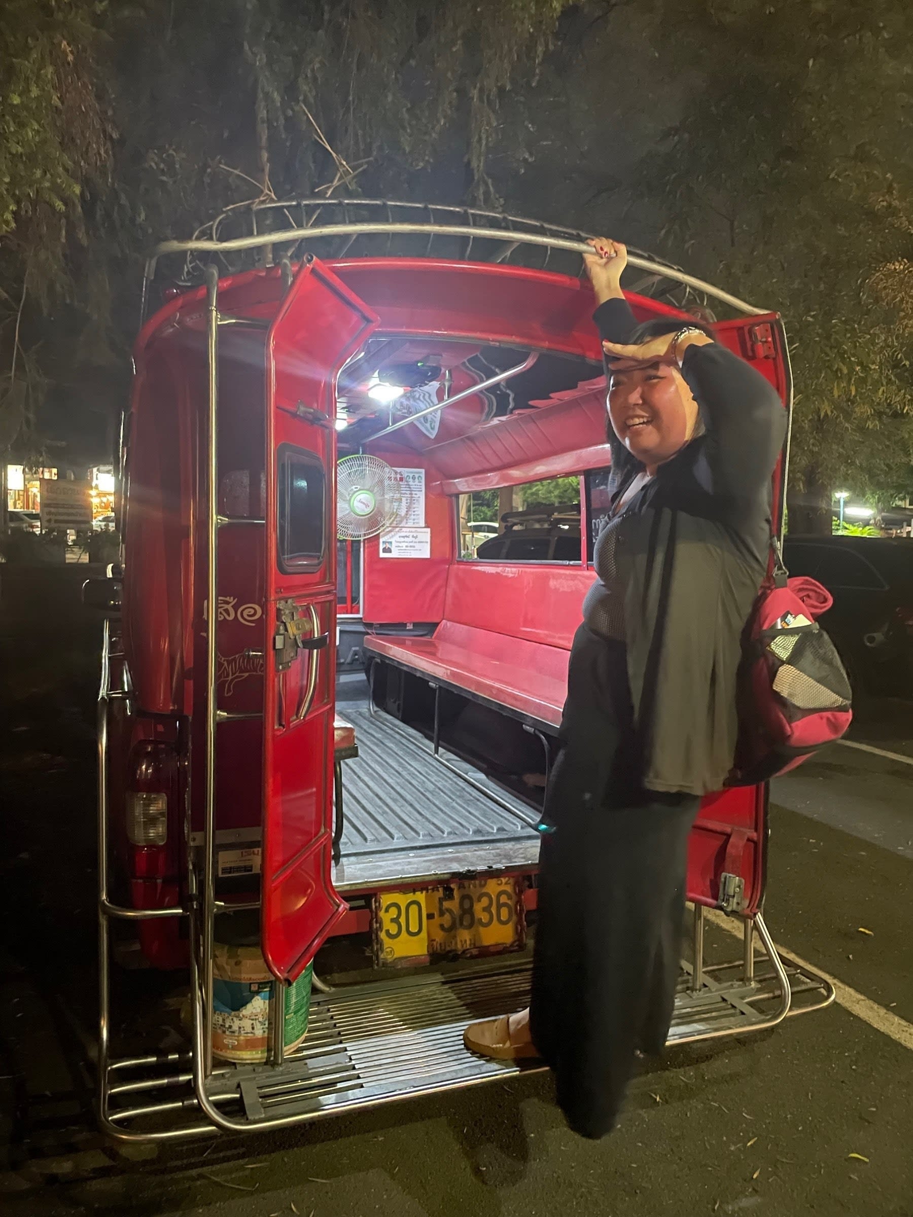 Chi hanging on the edge of a red songthaew, a Thailand public vehicle. She is mimicking the feeling of also hanging on the back end part of a Philippine jeepney, where commuters sometimes do this because the inside is already full.