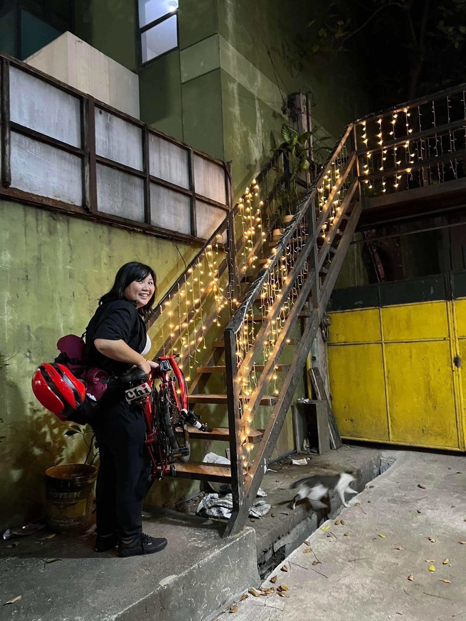 Chi about to carry her folded bike up some stairs that have Christmas or fairy lights draped on the railings, looking at the camera in an amused state. Her bike bag is in messenger bag mode, with her helmet strapped to the bag. A cat can also be seen blurry and running away from the stairs because it was initially chilling underneath it before Chi approached the area.