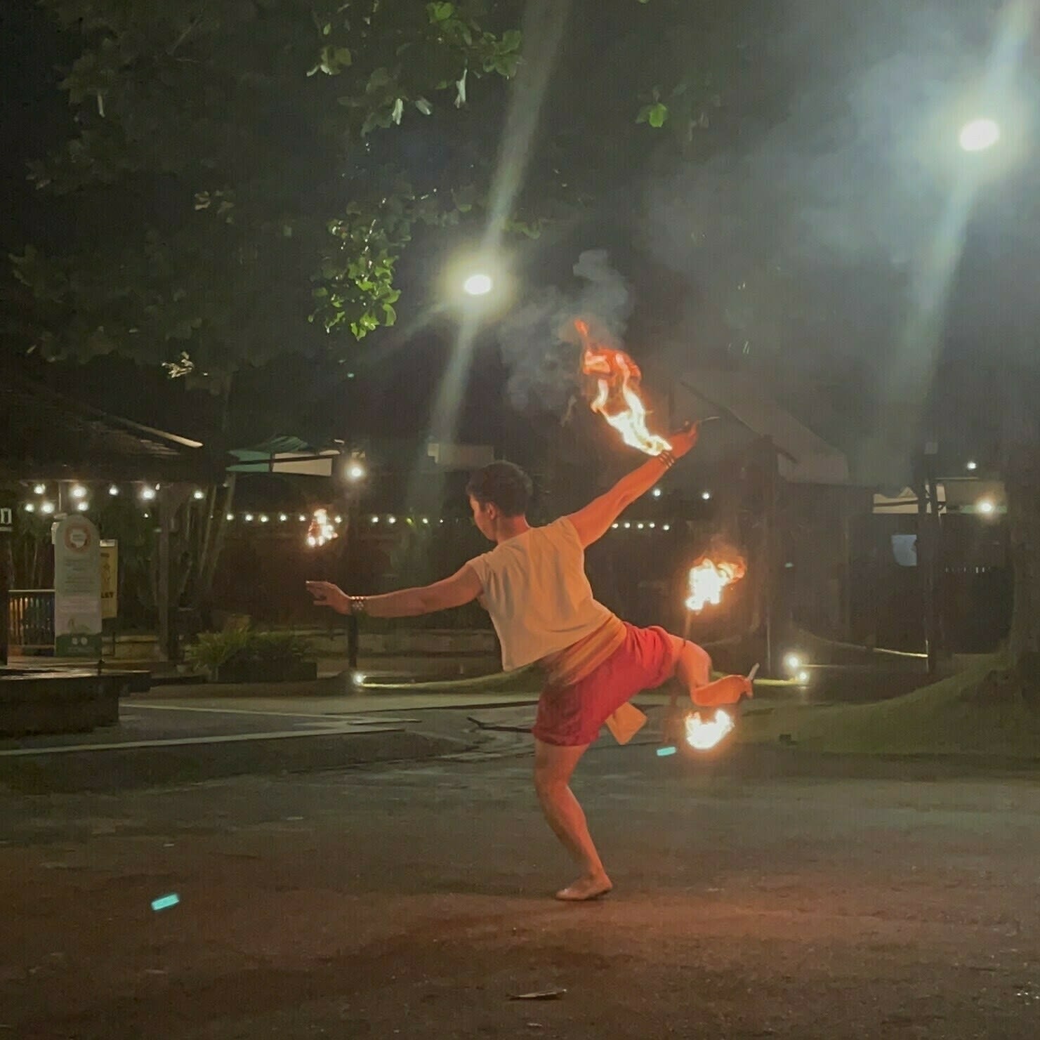 A Thai folk dancer holding onto 3 different lighted torches, 2 on their hands and one double-ended torch held by their right foot, dancing a traditional Thai dance. This performance was captured one evening in Chiang Mai, Thailand.