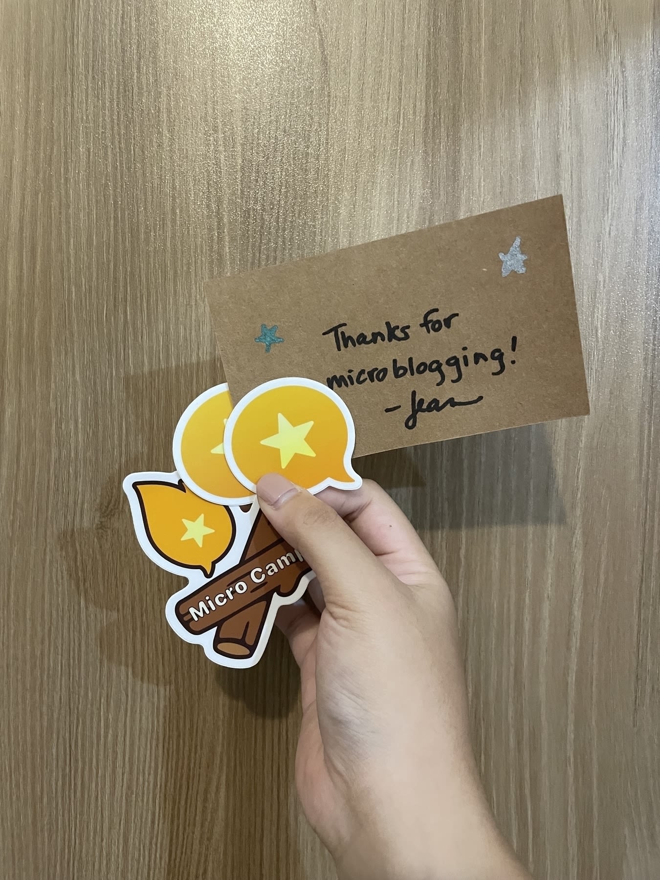 Chi’s hand holding up all the contents of the Micro.blog brown envelope sent to her, which includes 2 Micro.blog logo stickers, a Micro Camp campfire sticker, and a note from Jean which reads: “Thanks for microblogging!”
