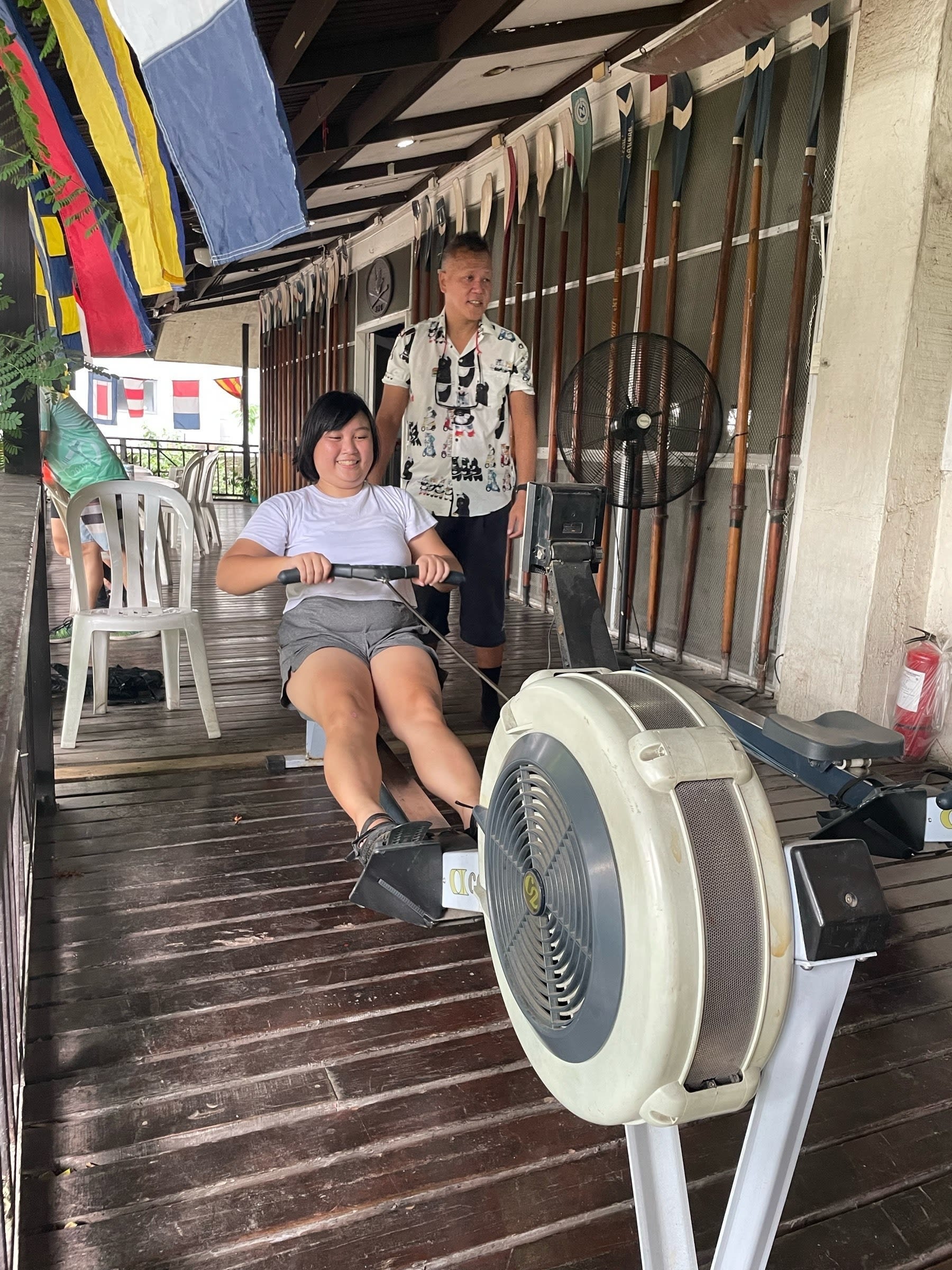 Chi seated on a rowing boat machine, with both her legs currently straightened and her hands on the rowing handlebars. A guide is seen to her left looking somewhere off-screen. They all are on the second floor of a wooden building, with wooden floorboards visible.
