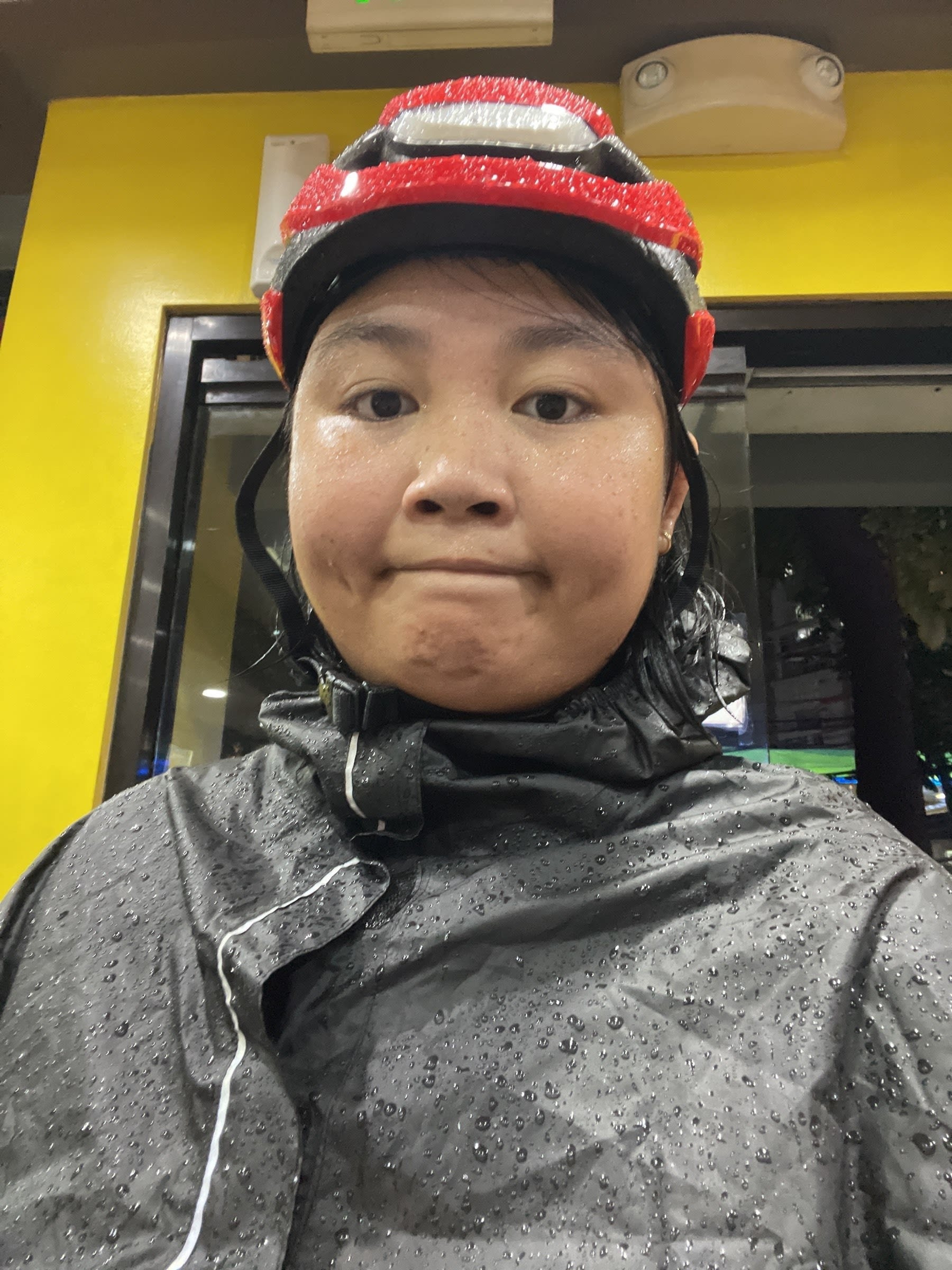 Selfie of Chi in her rain gear for her bike commute, wearing her red Lumos helmet and a black rain poncho. Both the poncho and her helmet are visibly drenched or have rain drops on them, and Chi’s making a dejected or kind of exhausted face.