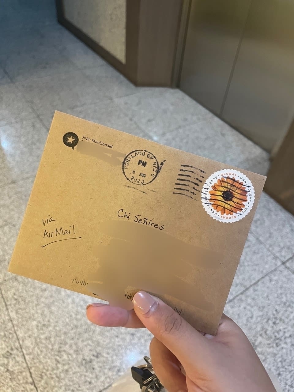 A brown envelope from Jean from Micro.blog, with address details obscured. There is a sunflower stamp on the upper right corner of the envelope, and a written underlined note on the lower left which reads, “via AirMail”.