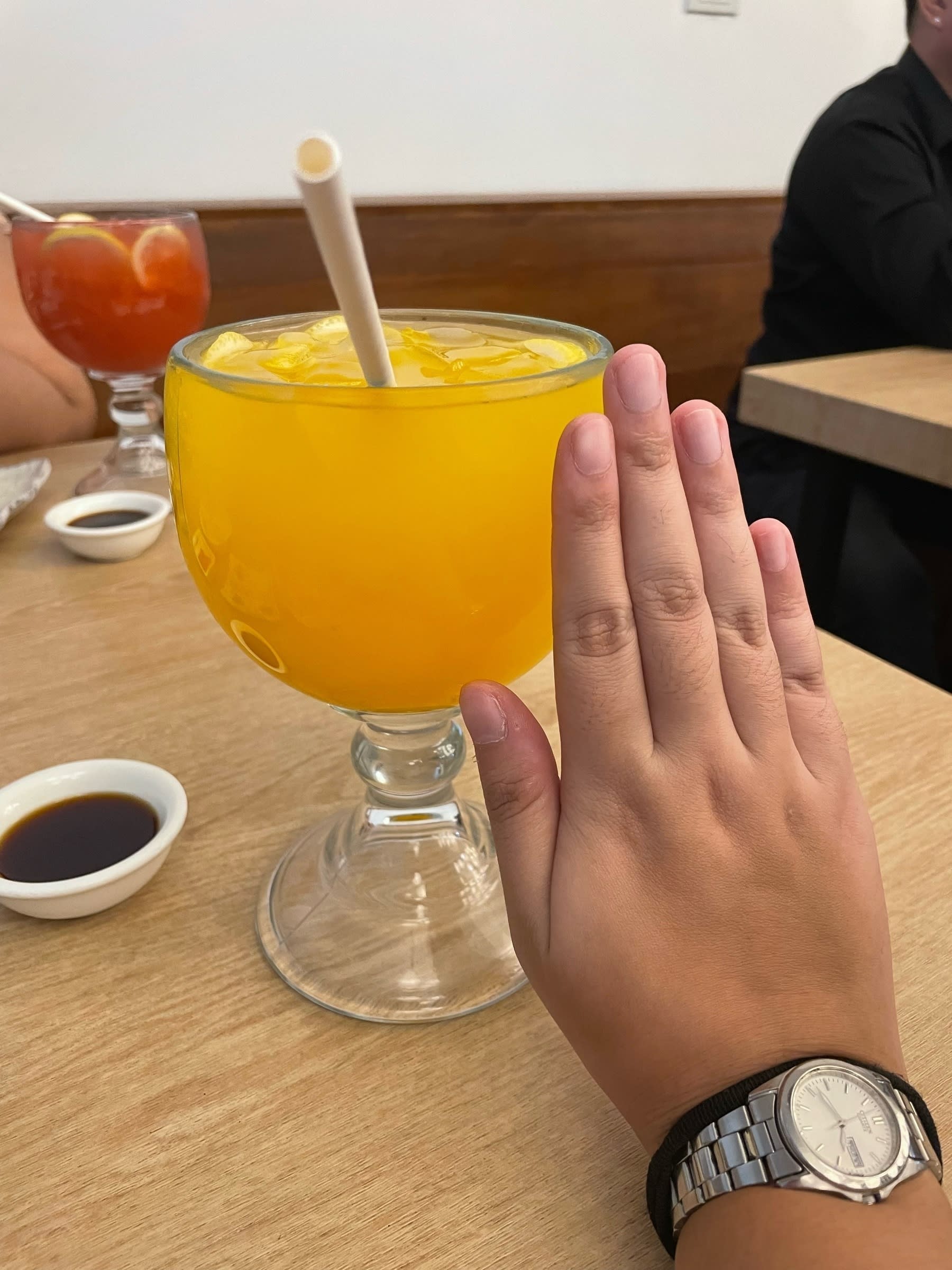 Chi’s glass filled with mango juice, with a really big glass. The size is as big and wide as her hand, which is positioned right next to the glass for comparison. In the background, her companion’s drink is also visible: a red drink, but Chi forgot what it was.