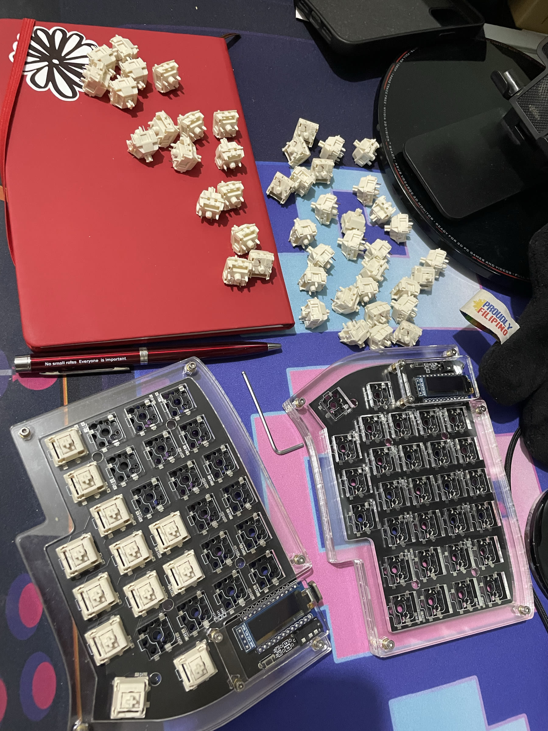 photo of Chi's desk with a bunch of keyboard switches scattered on top of notebook, and some keyboard switches also inserted already in the split keyboard frame. The printed circuit board can be seen through the split keyboard's acrylic case.