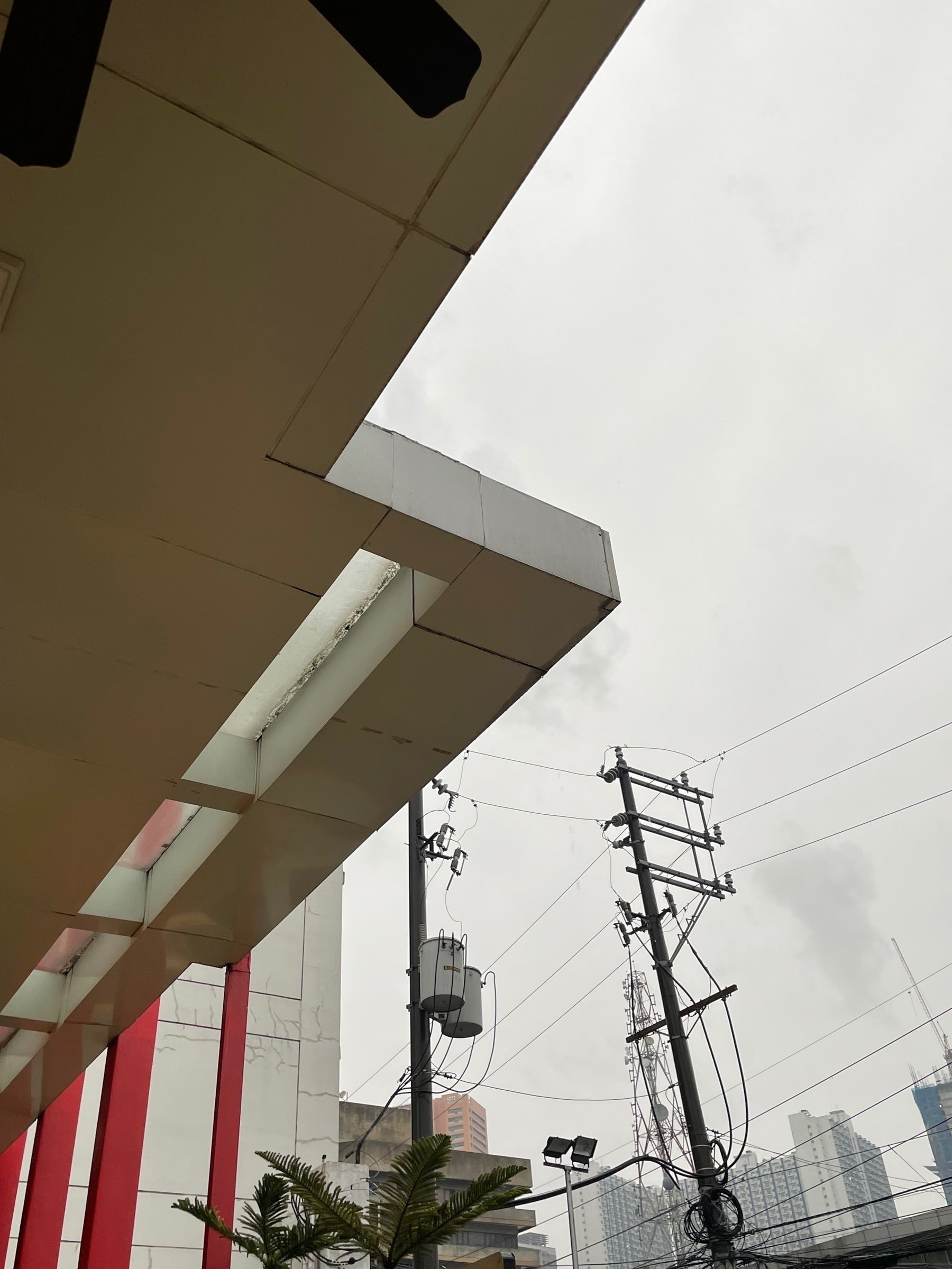The view underneath the roof of the McDonald’s branch Chi was eating breakfast at. There are a lot of corners on the roof, somewhat akin to a lightning bolt shape. Also seen in view are some above-ground electric lines, and further in the distance are some high-rise condominium buildings.