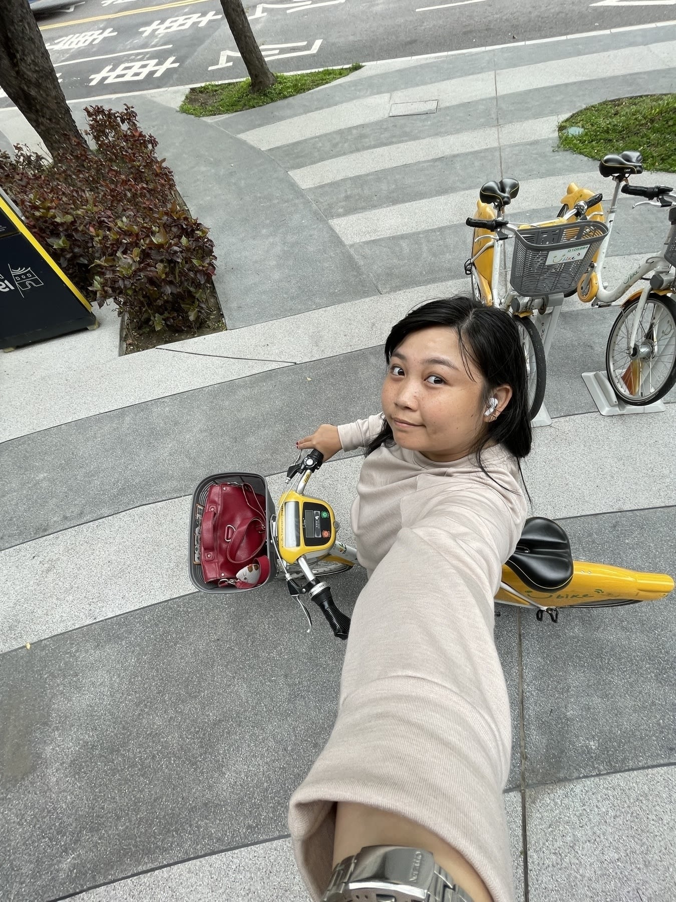 Wid angle selfie of Chi while on a rental YouBike. She’s wearing a cream-colored long-sleeved sweater, and her red leather bag is seen in the YouBike’s front basket. There are 2 other YouBikes parked behind her.