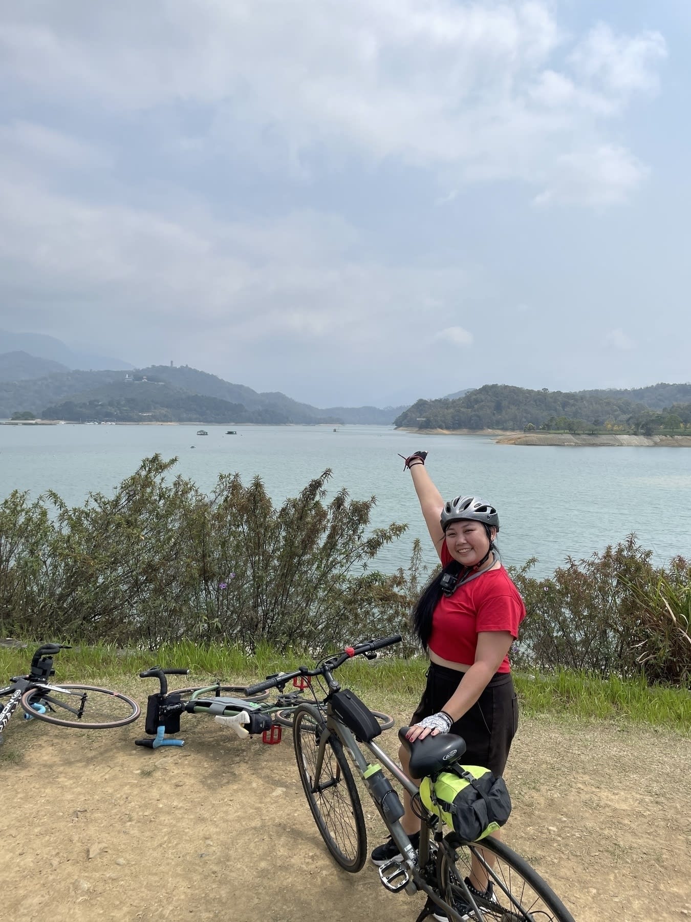 Chi posing with her right hand raised to gesture to her background, which is the Sun Moon Lake in Yuchi Township, Nantou County, Taiwan. She has the bike she borrowed from her friend parked in front of her, with the 2 other bikes of her friends laid on the ground just behind her. Chi is wearing a red crop top shirt and black shorts, as well as a bike helmet and fingerless gloves.