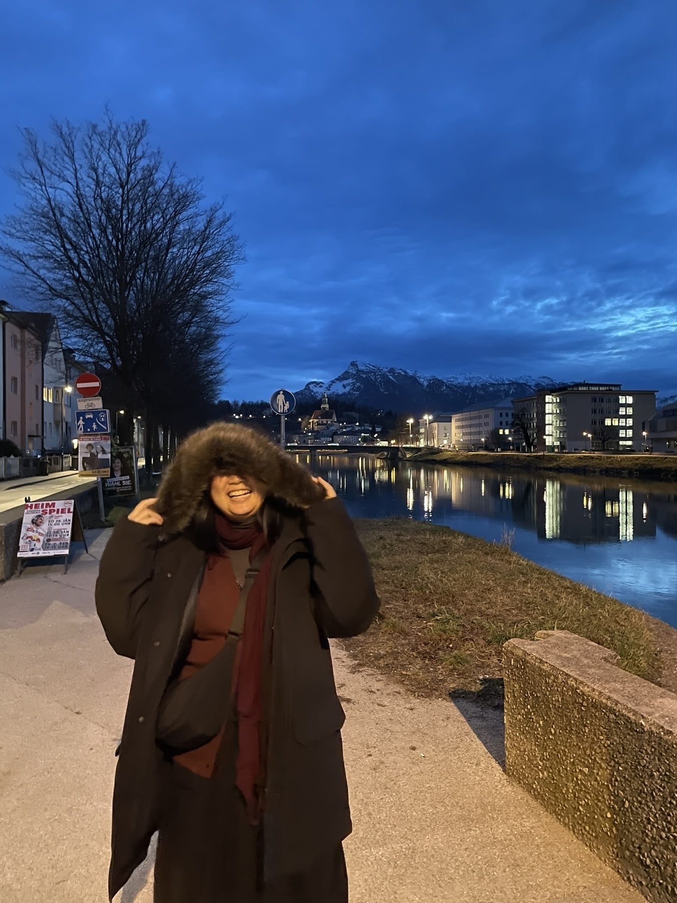 Chi is laughing while wearing her boyfriend’s black big winter jacket with a fluffy hoodie, because it looks like she’s being eaten by the jacket with the hoodie on top of her head. Behind her is the Salzach in Salzburg, Austria. The sky is dim, because it’s early evening.