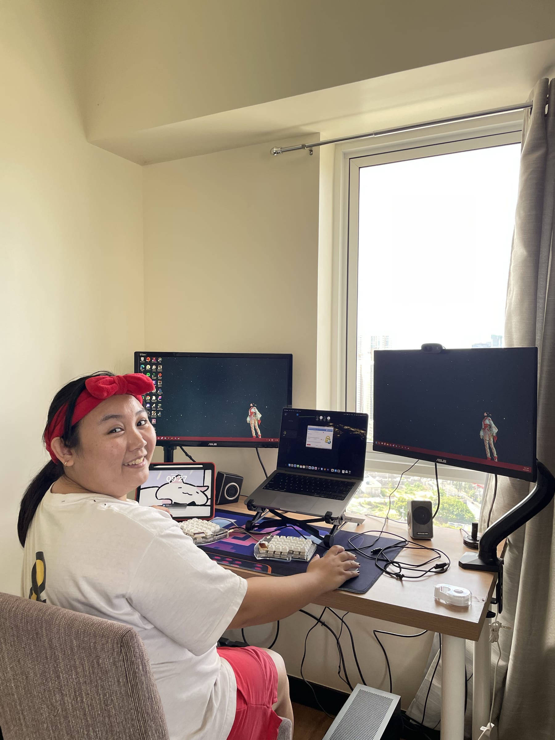 Chi looking at the camera and smiling while at her home workstation, consisting of two monitors mounted on monitor arms, an iPad propped up, and a Macbook Pro elevated with a laptop stand in the middle of her two monitors. On her desk is her split keyboard, her trackball mouse which Chi is holding, and a full deskmat. The chair Chi is sitting on is still a normal dining chair, not her computer chair. Her setup is facing the window, letting natural light in.