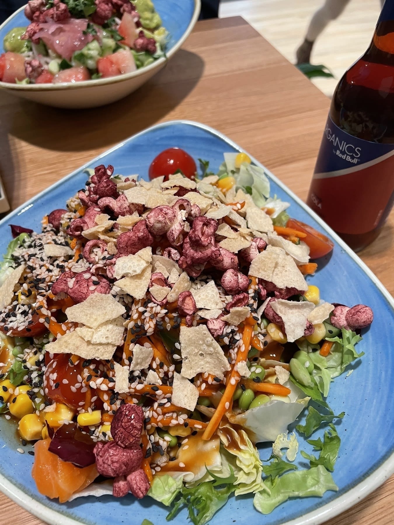 A poke bowl with hibiscus popcorn, rice flakes, corn, carrots, edamame, tomatoes, sesame seeds, and leafy greens all in a blue bowl.