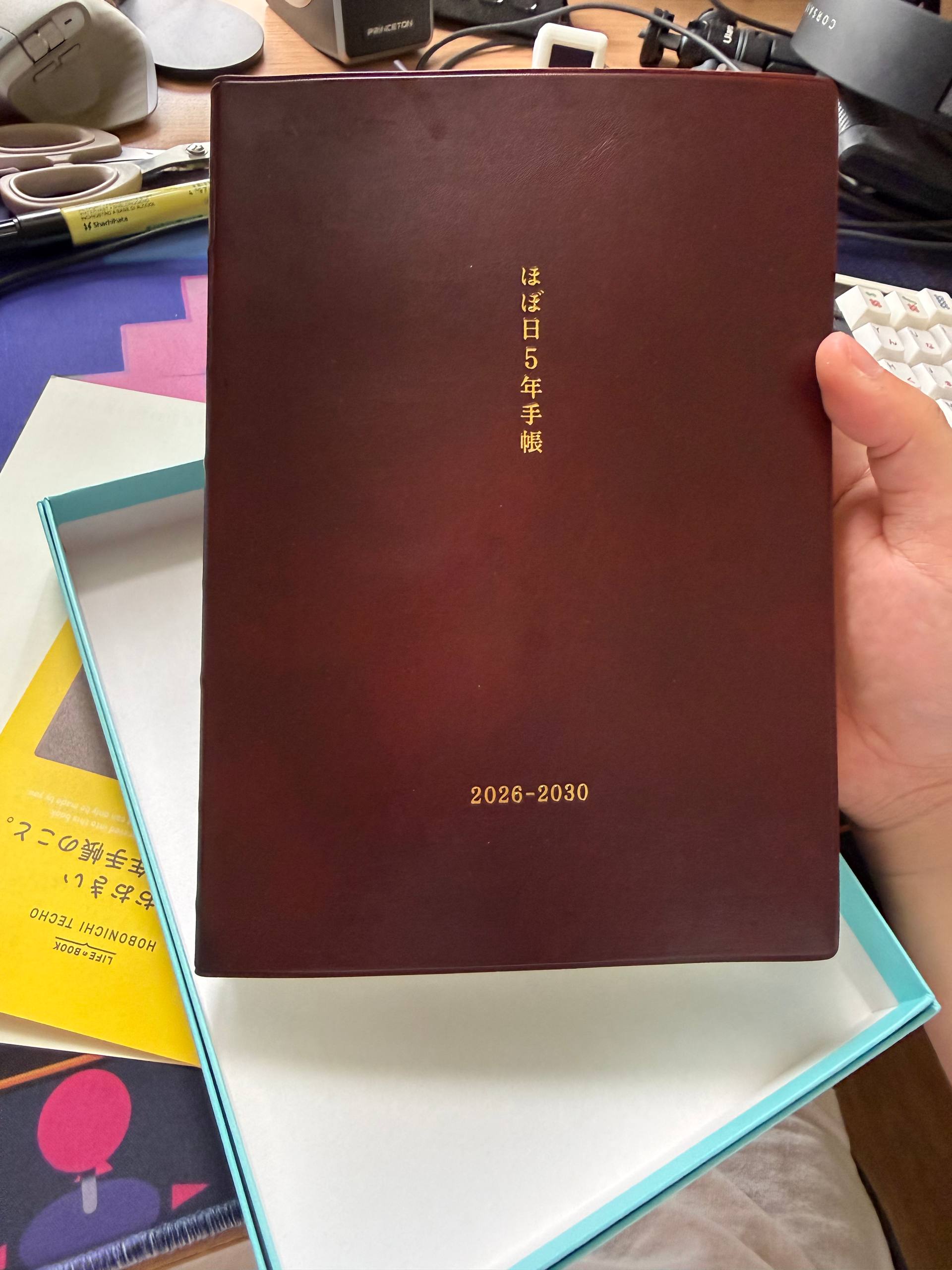 Chi holding up a hobonichi 5-year planner for the years 2026 to 2030. The brown leather cover has those labels embossed in gold colored Japanese characters.