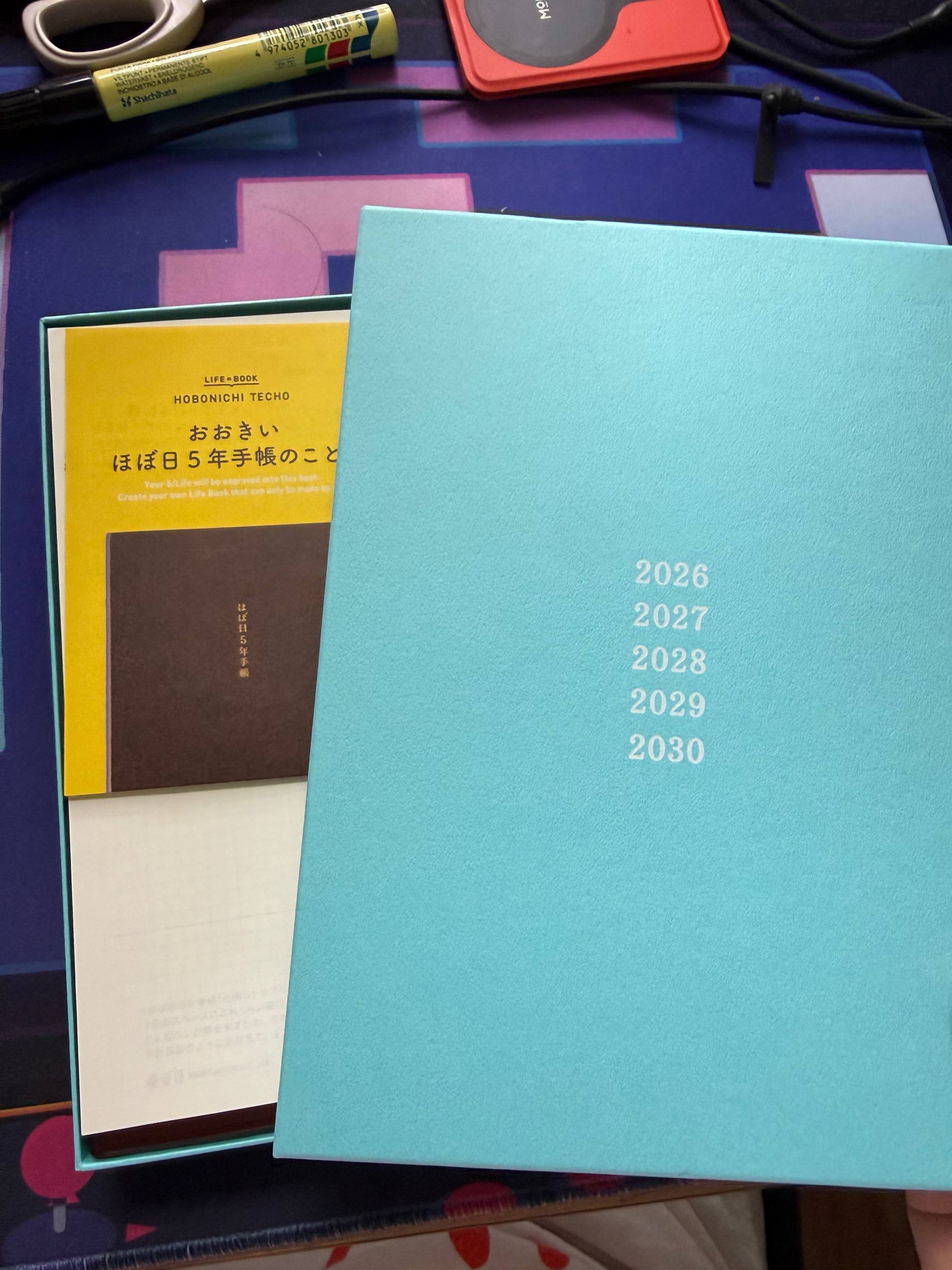 A partially opened box for the hobonichi techo life book, with the years 2026 to 2030 printed on the teal box cover set to the right. Inside the box is a paper instruction card with writing in Japanese, reading as "about the big hobonichi 5-year planner".