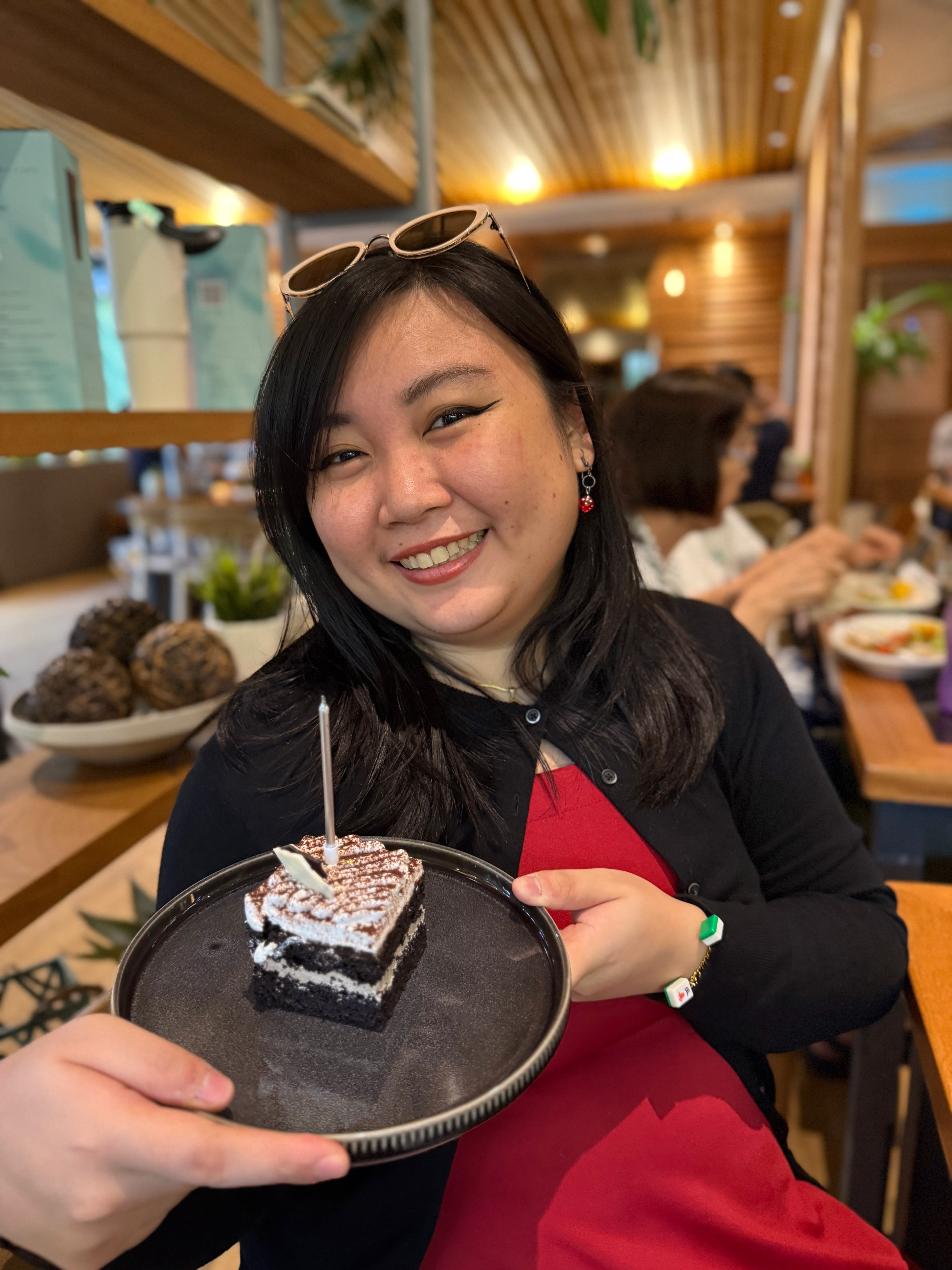 Chi smiling and holding a plate with a slice of chocolate cake with a silver candle. She’s wearing red dice earrings, a black cardigan, a red dress, and her mahjong trinket bracelet. She also has her shades on her head.