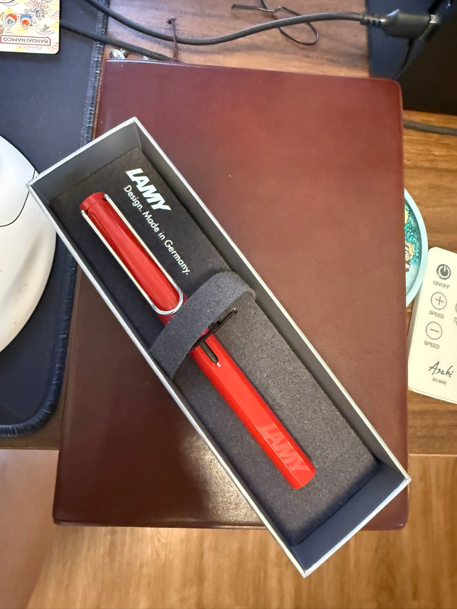 A red Lamy Safari fountain pen still in its box, with the Lamy logo and the tagline, “Design. Made in Germany.” printed on the upper left portion of the box that is placed diagonally on Chi’s brown hobonichi planner.