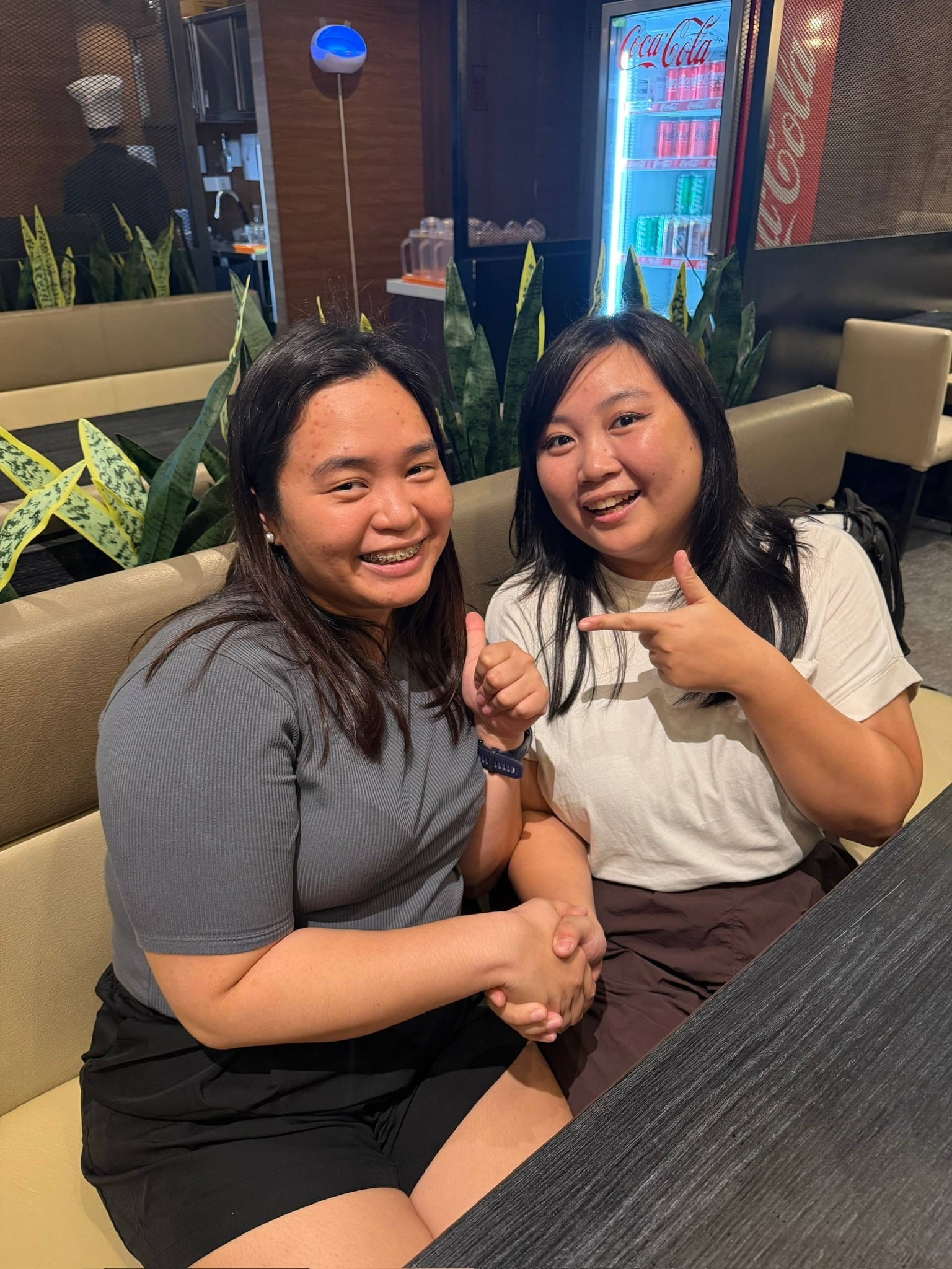 Jas and Chi seated in a restaurant, smiling at the photo. Jas is holding a thumbs up gesture while shaking hands with Chi, while Chi has her left hand pointing to Jas.