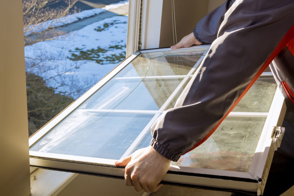 A person wearing a gray and red jacket is installing or removing a window pane from a window frame on a snowy day.