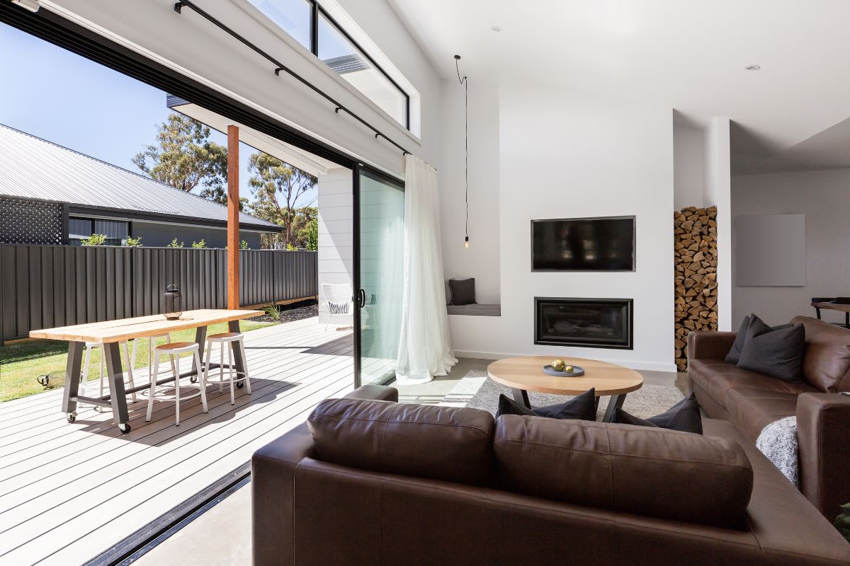 Modern living room with brown leather couches, a round coffee table, and a wall-mounted TV. The space opens to an outdoor patio via sliding glass doors. Firewood is stacked next to the fireplace.