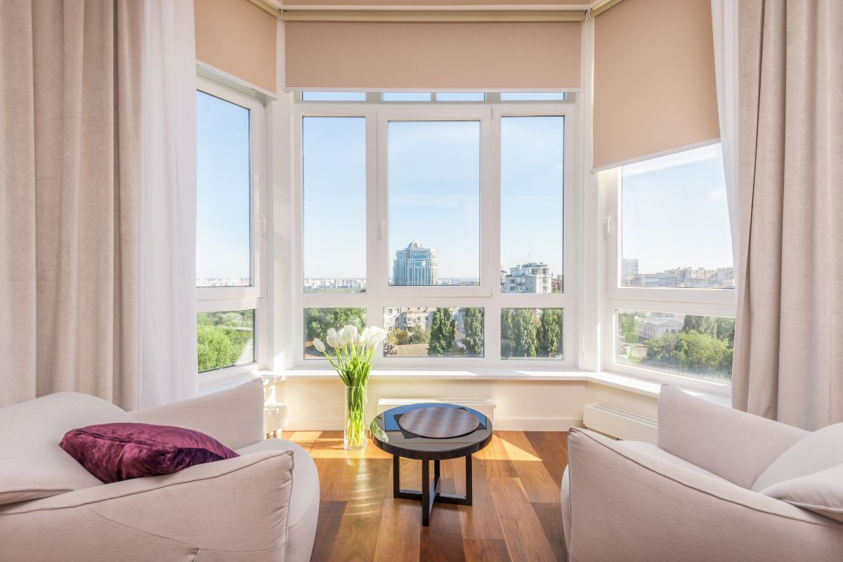 A bright room with large windows, embracing the best types of windows for homes, features two beige armchairs, a small round table with a vase of flowers, and an impressive cityscape view in the background.