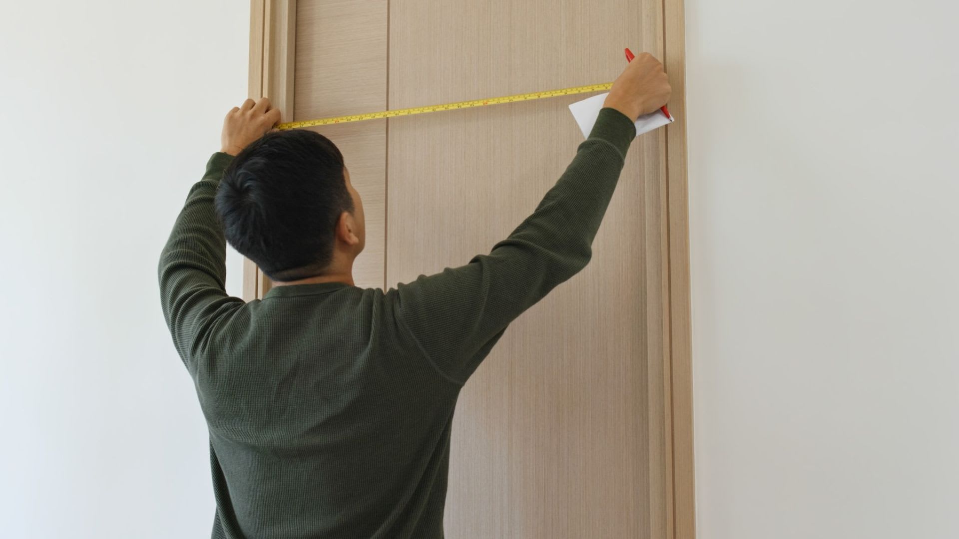 A person demonstrates how to measure a door by using a yellow tape measure to check the width of a wooden door, holding a pen and paper in one hand.