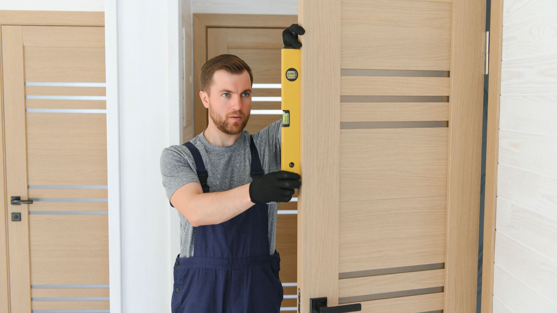 A man in work overalls and gloves uses a spirit level to check the alignment of a wooden door indoors, demonstrating precise door measurements for ensuring the perfect fit.