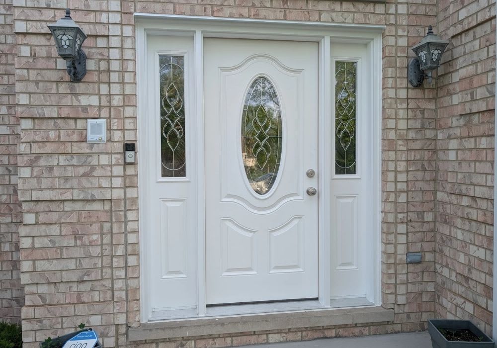 White front door with an oval glass window and two narrow glass panels on each side, set in a light brick exterior. Two outdoor wall lights are mounted on either side of the door.