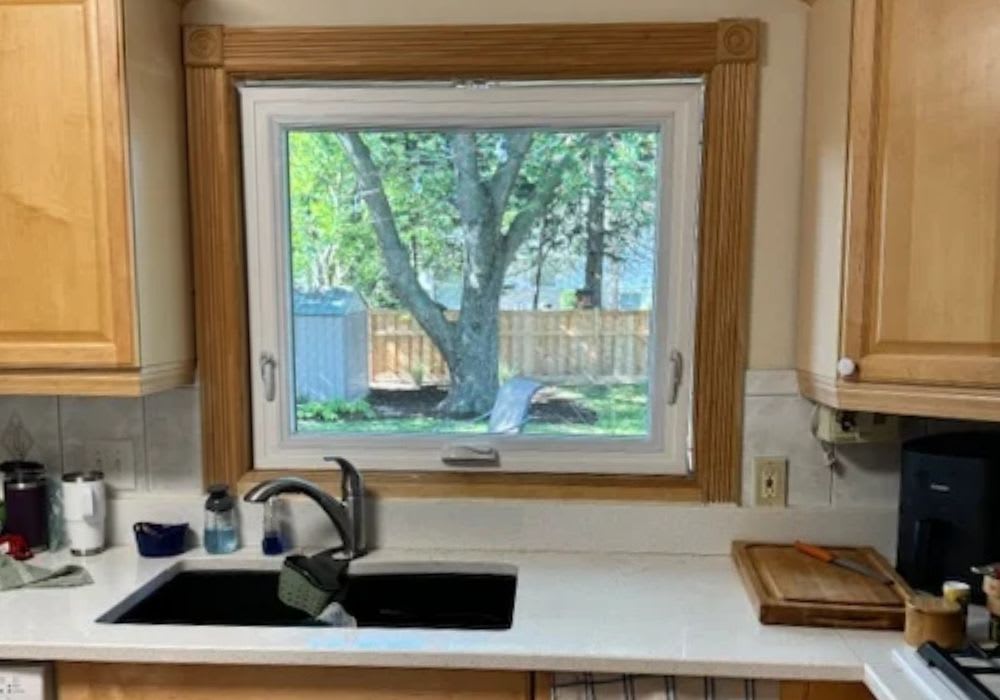 A kitchen sink with a faucet is centered under a window that looks out onto a backyard with a tree, a shed, and a wooden fence.