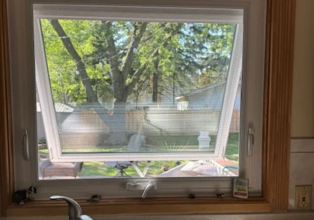 A kitchen window is tilted open inward above a sink, showing a green backyard with trees and a wooden fence outside.