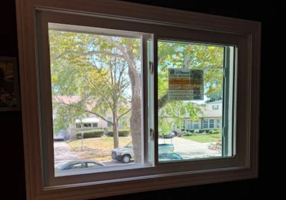 A view through a double-pane window showing a residential street with trees, parked cars, and houses outside. A temporary sticker is attached to the window.